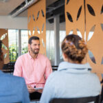 Back view of employees listening to young consultant. Colleagues sitting on chairs and listening to smiling creative director. Business meeting concept